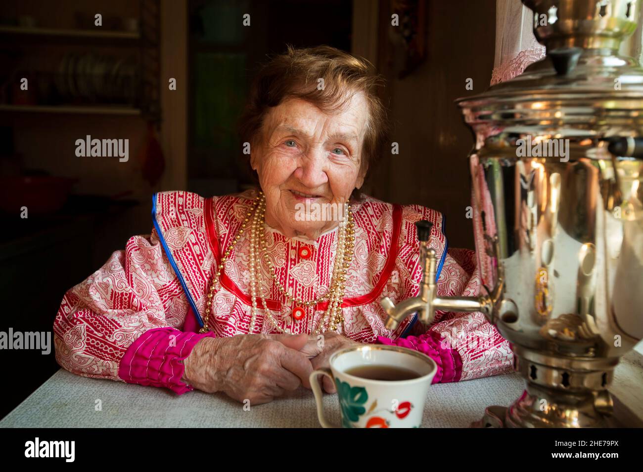 Une femme âgée en robe nationale russe est assise en train de boire du thé avec un samovar russe dans sa maison. Banque D'Images
