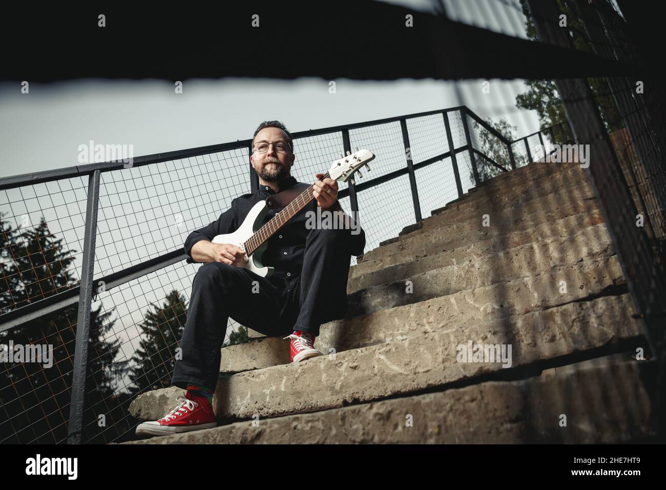 Portrait d'un guitariste mâle avec guitare électrique portant des vêtements noirs et des snegerers rouges assis sur un escalier en zone urbaine Banque D'Images