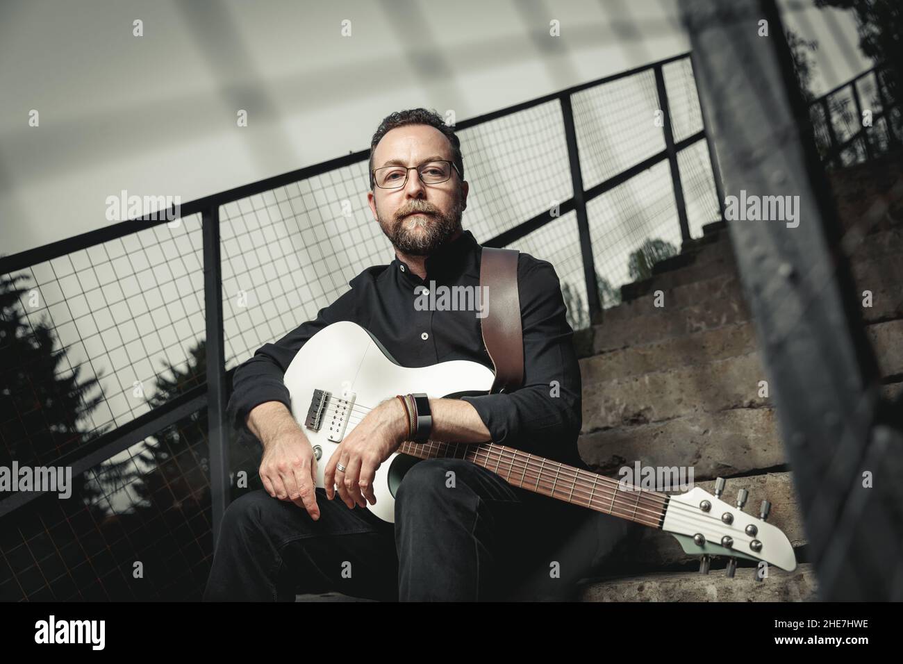 Portrait d'un guitariste mâle avec guitare électrique portant des vêtements noirs et des snegerers rouges assis sur un escalier en zone urbaine Banque D'Images