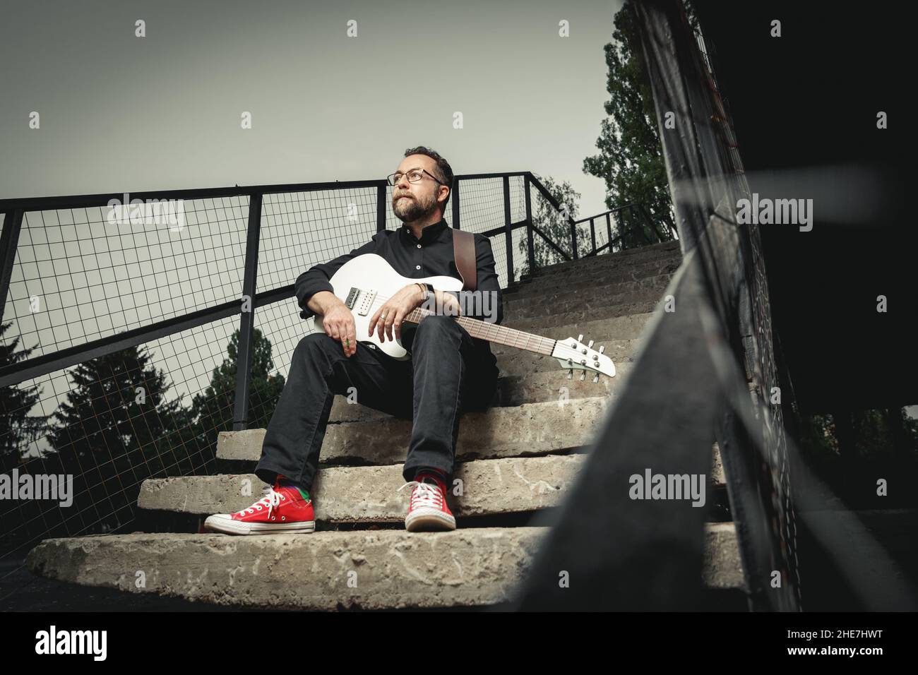 Portrait d'un guitariste mâle avec guitare électrique portant des vêtements noirs et des baskets rouges assis sur un escalier en zone urbaine Banque D'Images