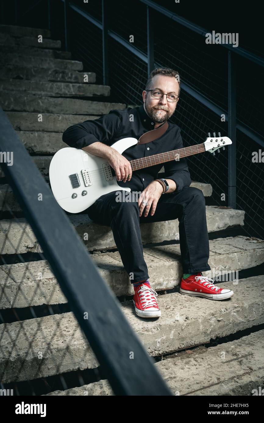 Portrait d'un guitariste mâle avec guitare électrique portant des vêtements noirs et des baskets rouges assis sur un escalier en zone urbaine Banque D'Images