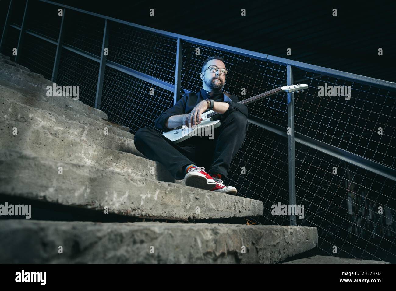 Portrait d'un guitariste mâle avec guitare électrique portant des vêtements noirs et des baskets rouges assis sur un escalier en zone urbaine Banque D'Images