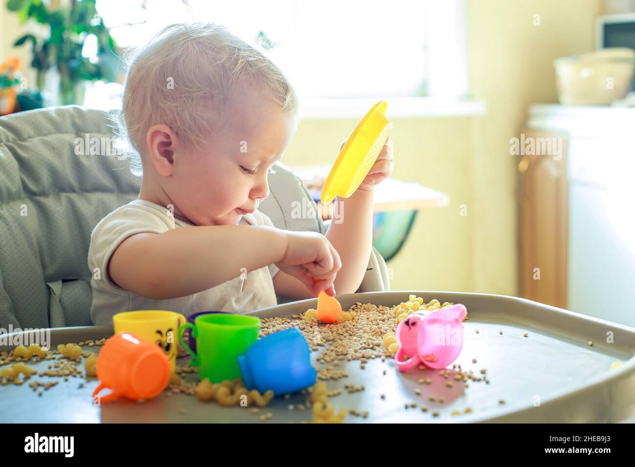 petit enfant assis sur une chaise d'alimentation et joue avec des ustensiles de jouet et du grain. développement de compétences en moteur fin chez les enfants jusqu'à l'année. Développement précoce Banque D'Images