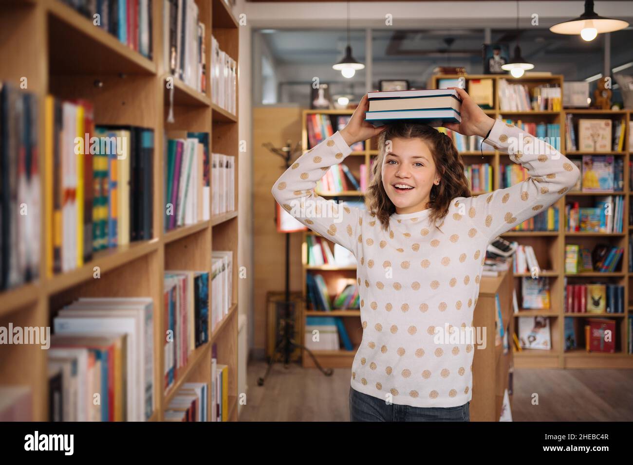 Fille de l'adolescence parmi une pile de livres.Une jeune fille tenant des livres sur la tête avec des étagères en arrière-plan.Elle est entourée de piles de livres.Réservez le jour. Banque D'Images
