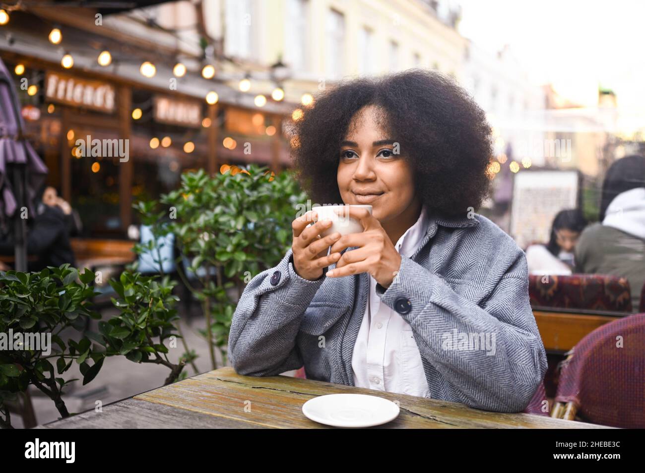 Mode Street style portrait de jeune beauté naturelle attrayante femme afro-américaine avec des cheveux afro dans une veste en tweed posant à l'extérieur dans un café-terrasse.Bonne dame avec une tasse de café dans la grande ville Banque D'Images