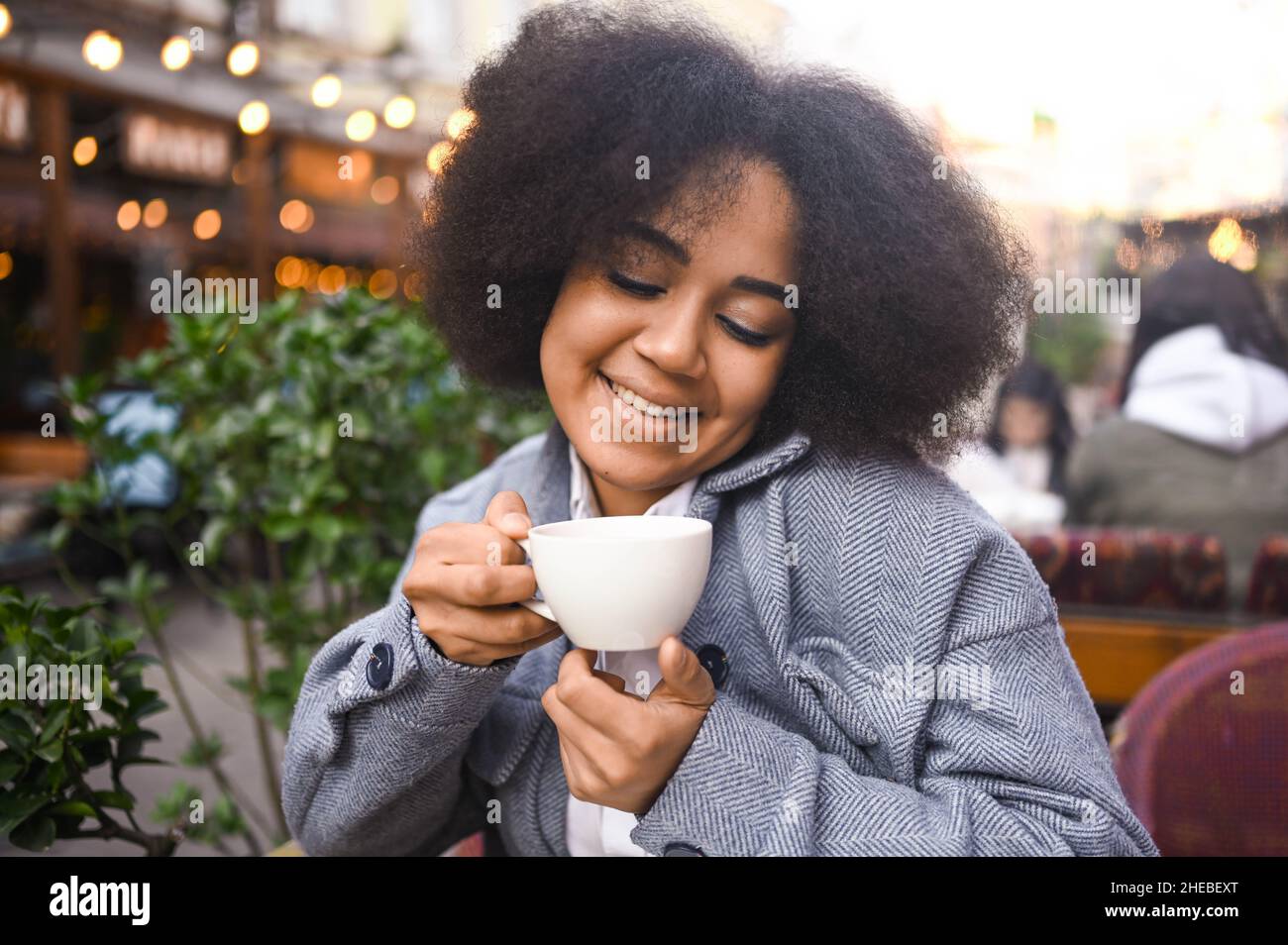 Mode Street style portrait de jeune beauté naturelle attrayante femme afro-américaine avec des cheveux afro dans une veste en tweed posant à l'extérieur dans un café-terrasse.Bonne dame avec une tasse de café dans la grande ville Banque D'Images
