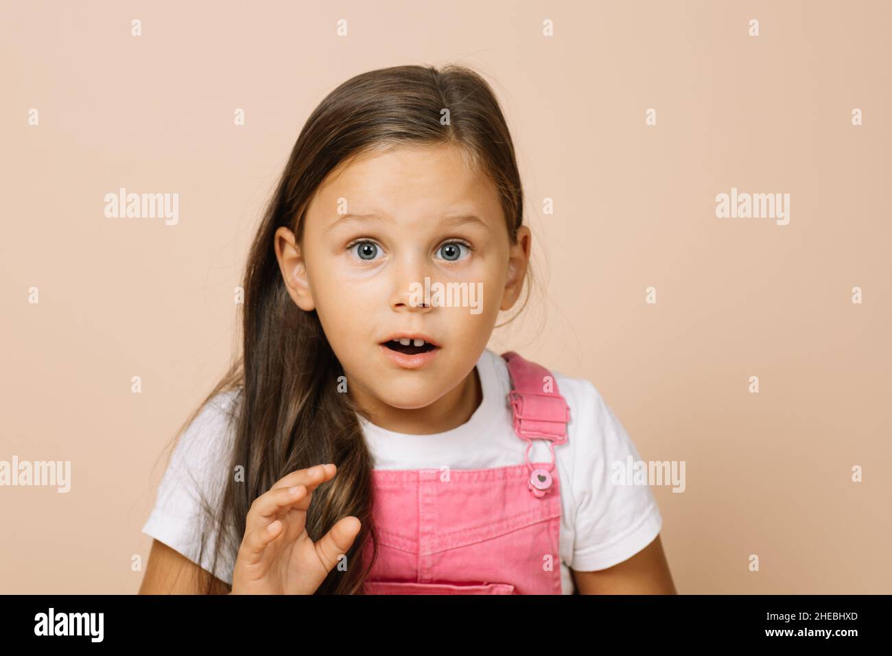 Portrait d'un enfant avec des yeux ronds surpris, une bouche légèrement ouverte et une main levée regardant l'appareil photo portant une combinaison rose vif et un t-shirt blanc Banque D'Images