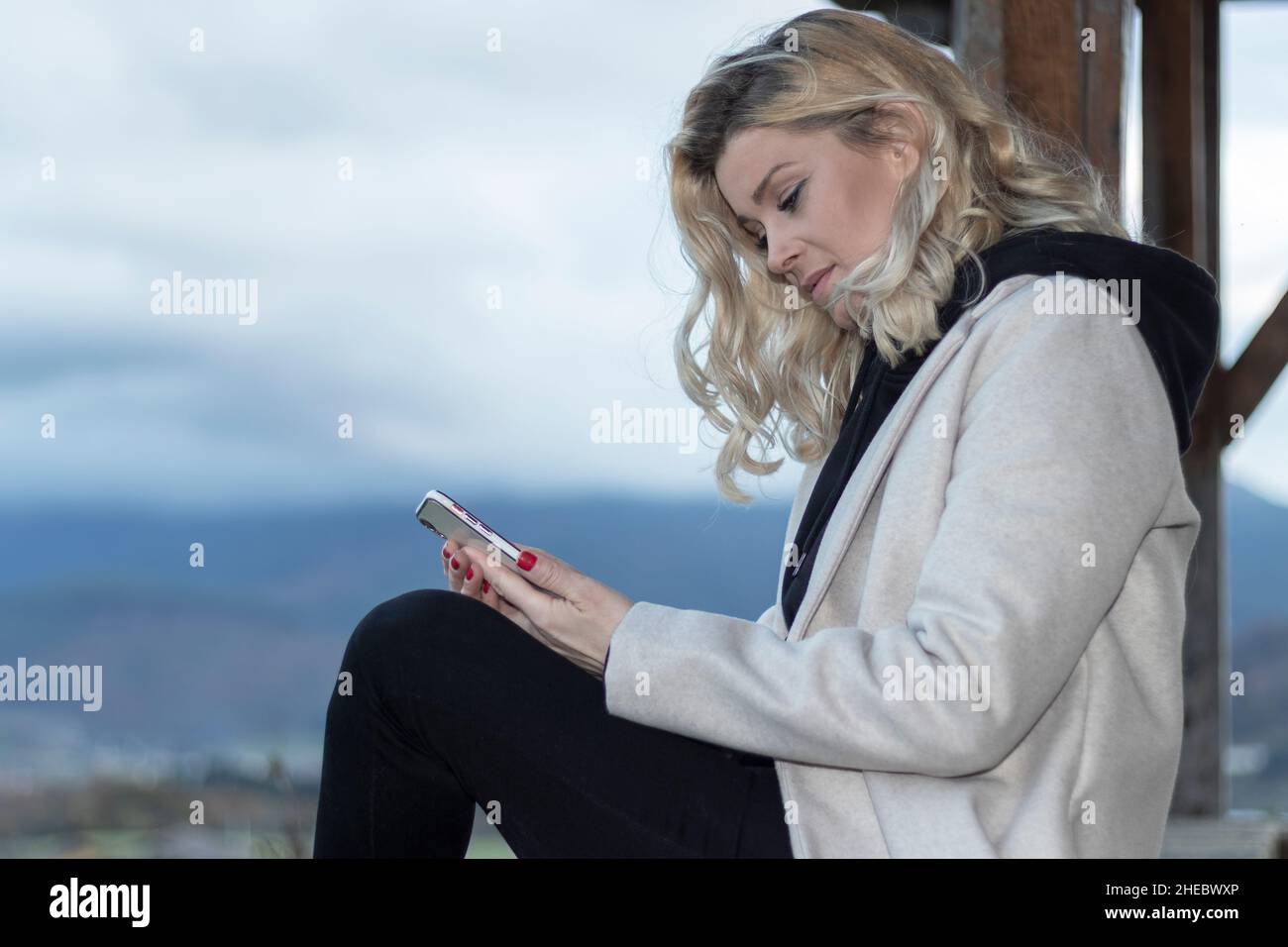 blonde regardant le mobile assis sur un mur au coucher du soleil Banque D'Images