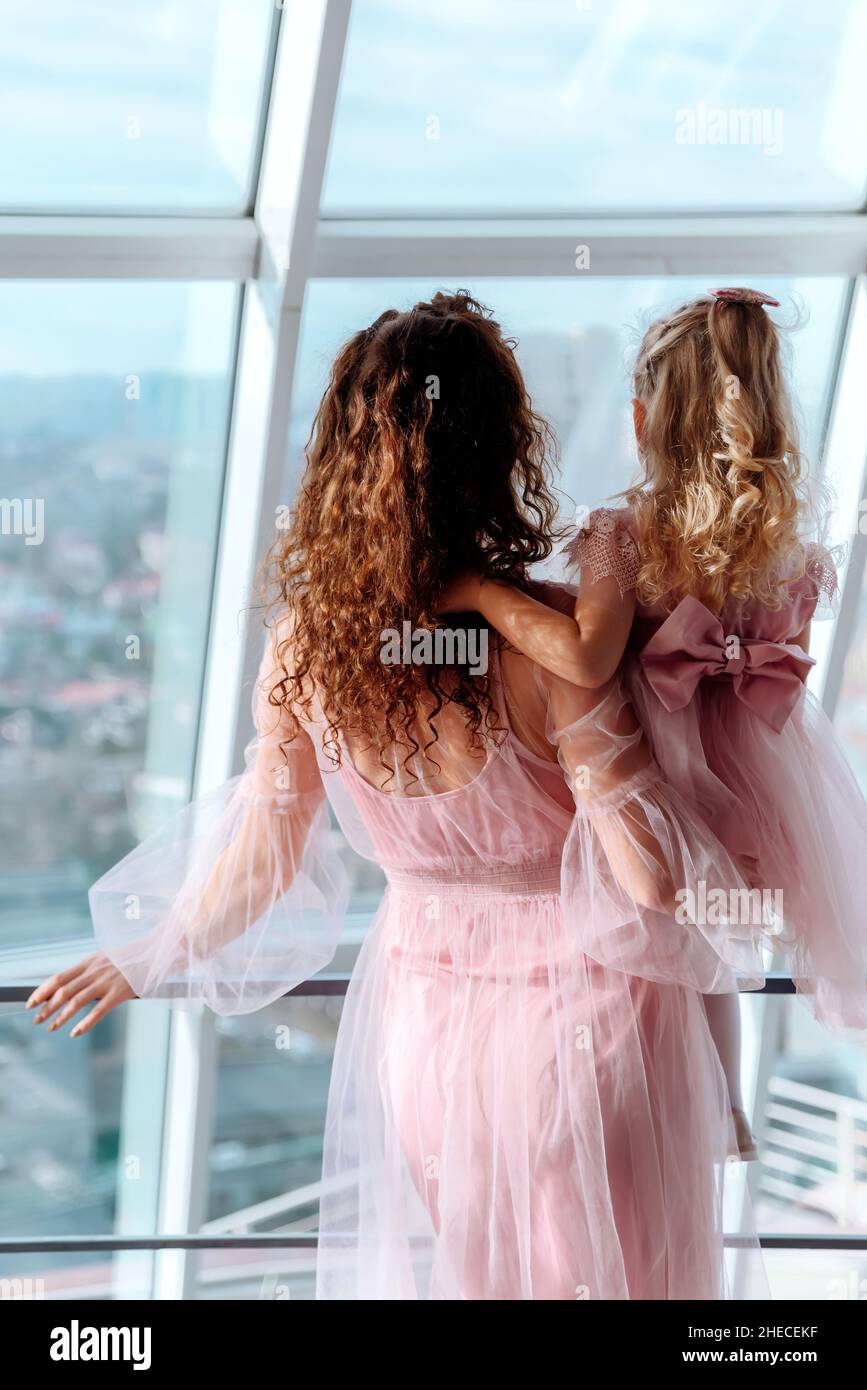 Une mère avec une petite fille dans ses bras portant des robes roses  identiques se trouve à une grande fenêtre panoramique.Contenu de maternité,  concept de fête des mères.Vue arrière, prise de vue