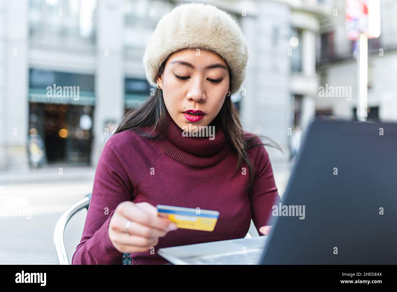 Concentrée chinoise féminine avec carte de crédit faisant l'achat en ligne sur ordinateur portable tout en étant assis sur la terrasse du café dans la ville moderne Banque D'Images