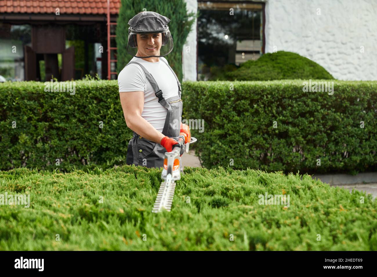 Beau caucasien homme en uniforme, défenseur du visage et gants de protection coupant des buissons trop grands sur la cour arrière.Jardinier compétent utilisant un taille-haie à essence pour le travail en extérieur. Banque D'Images