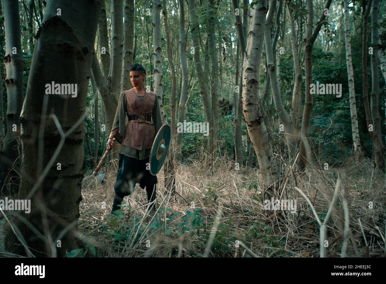 Femme sérieuse avec du sang sur le visage en costume viking traditionnel debout dans la forêt avec la hache à la main Banque D'Images