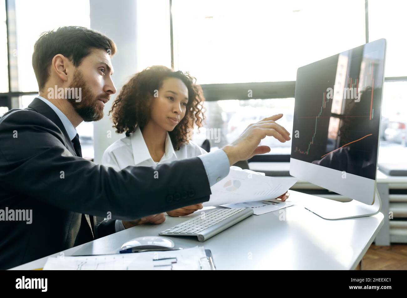 Homme caucasien et femme afro-américaine, commerçants de crypto, assis dans un bureau, en utilisant un ordinateur, en regardant des graphiques, en analysant le marché de crypto, en planifiant une stratégie pour de bons profits Banque D'Images