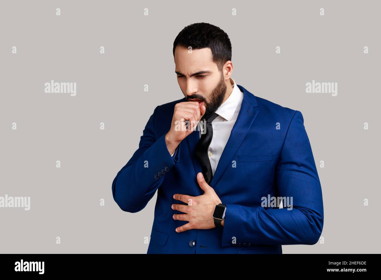 Portrait d'un homme barbu malsain toussant, attrape froid, ayant une température élevée, ayant un symptôme de grippe, portant un costume de style officiel.Prise de vue en studio isolée sur fond gris. Banque D'Images