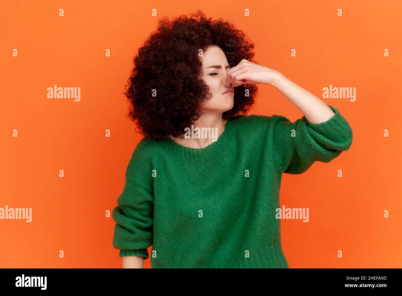 Portrait d'une jeune femme adulte avec une coiffure afro portant un chandail vert de style décontracté tenant la respiration avec les doigts sur le nez, odeur piquant, fart.Studio d'intérieur isolé sur fond orange. Banque D'Images