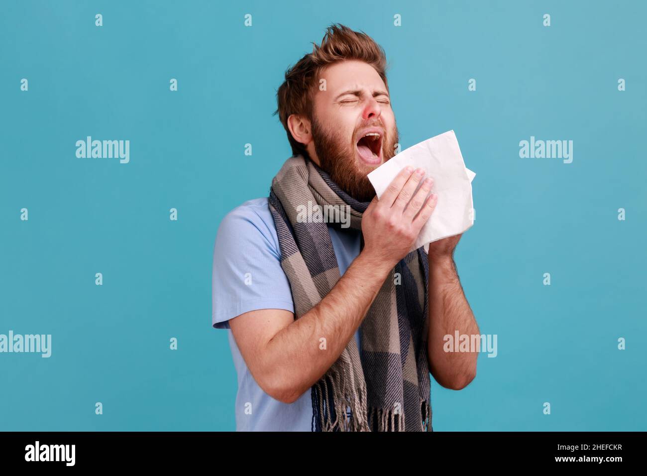 Épidémie de virus, épidémie de grippe.Homme enveloppé d'un foulard éternuant dans une serviette, nettoyant le nez et la toux, souffrant de symptômes grippaux, fièvre.Studio d'intérieur isolé sur fond bleu. Banque D'Images