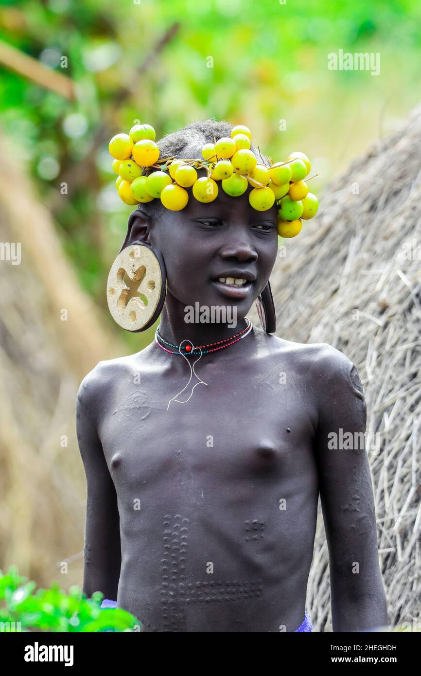 Omo River Valley, Éthiopie - 29 novembre 2020 : portrait d'un adolescent africain avec de grandes boucles d'oreilles traditionnelles en bois et une couronne de fleurs jaunes sèches Banque D'Images
