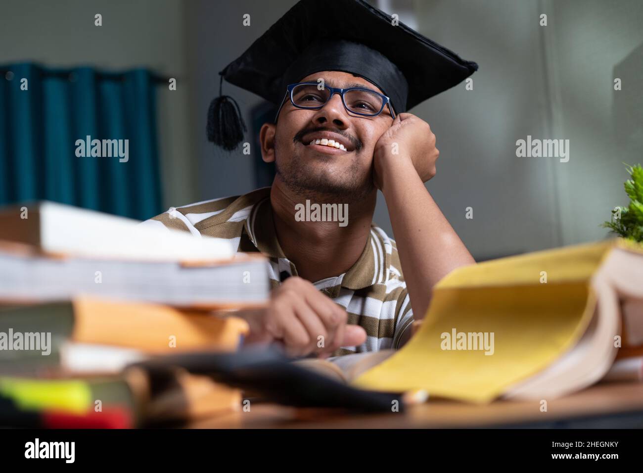 jeune étudiant rêvant de la remise des diplômes tout en studant le livre ou en se préparant à l'examen à la maison - concept de rêverie et futur objectif de carrière et Banque D'Images