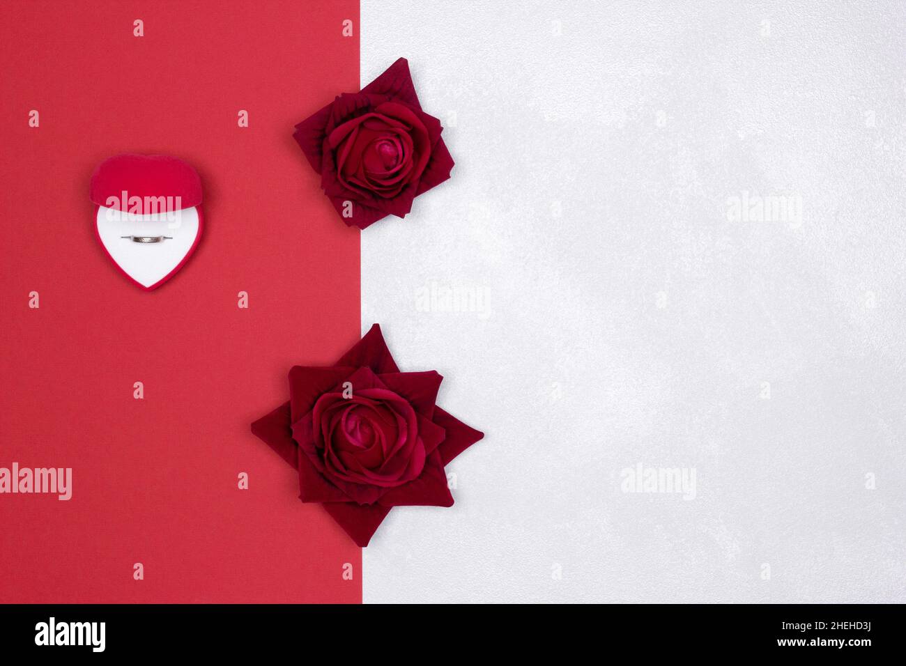 Boîte cadeau ouverte en velours rouge en forme de coeur avec anneau de mariage à l'intérieur et fleurs roses sur fond texturé rouge et blanc argenté.Copier l'espace Banque D'Images