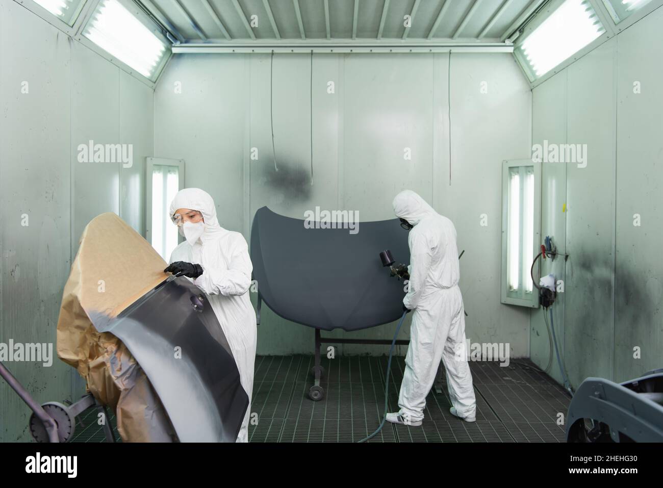 Jeune femme de travail en costume de noisette et masque de protection debout près de la partie de voiture tandis que collègue utilisant l'aérographe dans le garage Banque D'Images