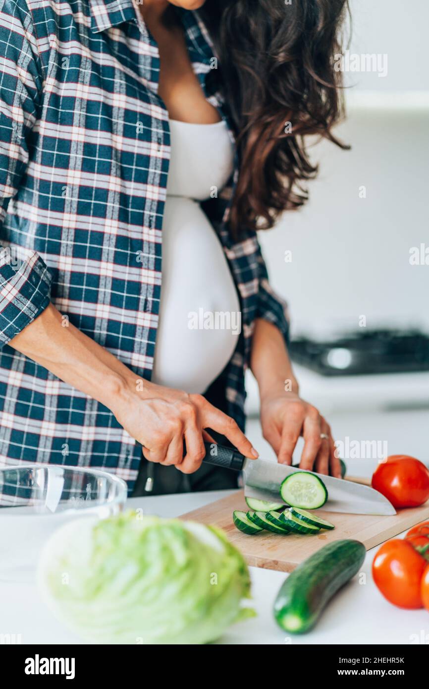 Une femme prégnante méconnue prépare une salade de concombre sur une planche à découper en bois en mettant des légumes frais et des fruits sur une table dans la cuisine.Grossesse, ma Banque D'Images