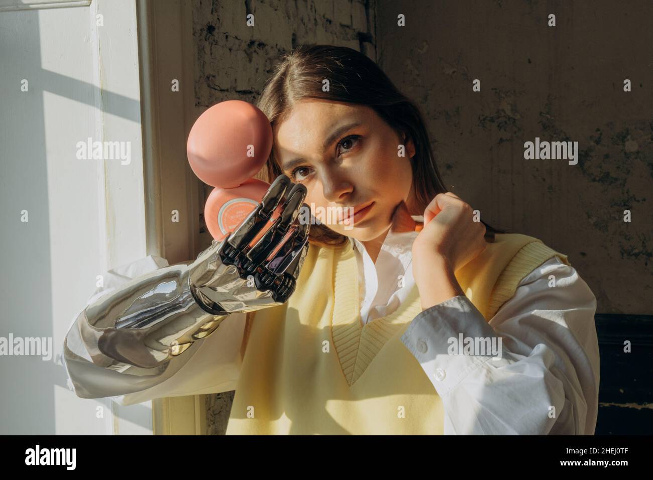 Belle femme avec un bras prothétique bionique applique le maquillage avec une brosse debout à la fenêtre un jour ensoleillé et regarde dans un petit miroir. Banque D'Images