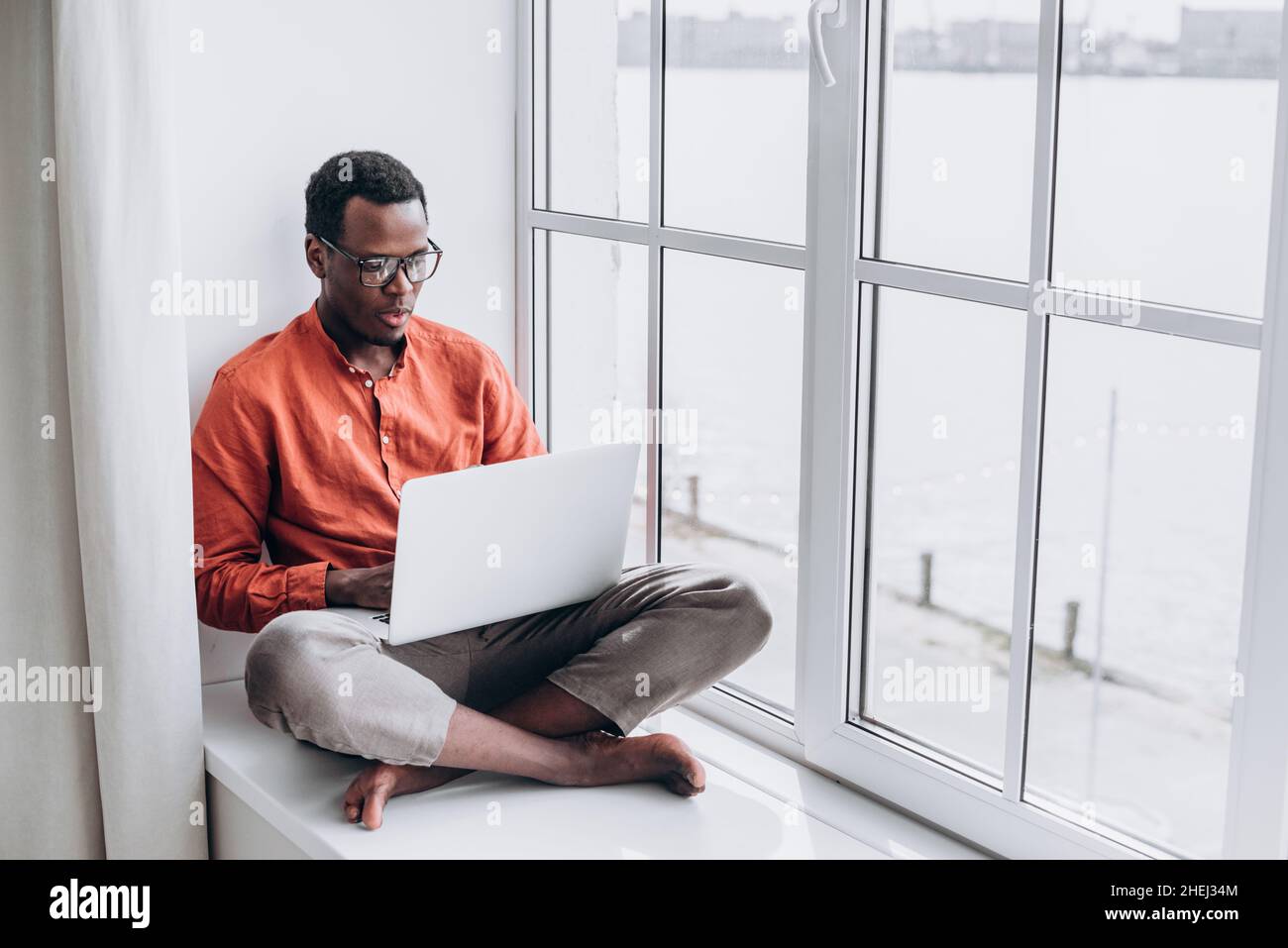 Un homme afro-américain séduisant en vêtements et lunettes décontractés surfe sur Internet sur un ordinateur portable moderne assis sur le seuil de la fenêtre à la maison Banque D'Images