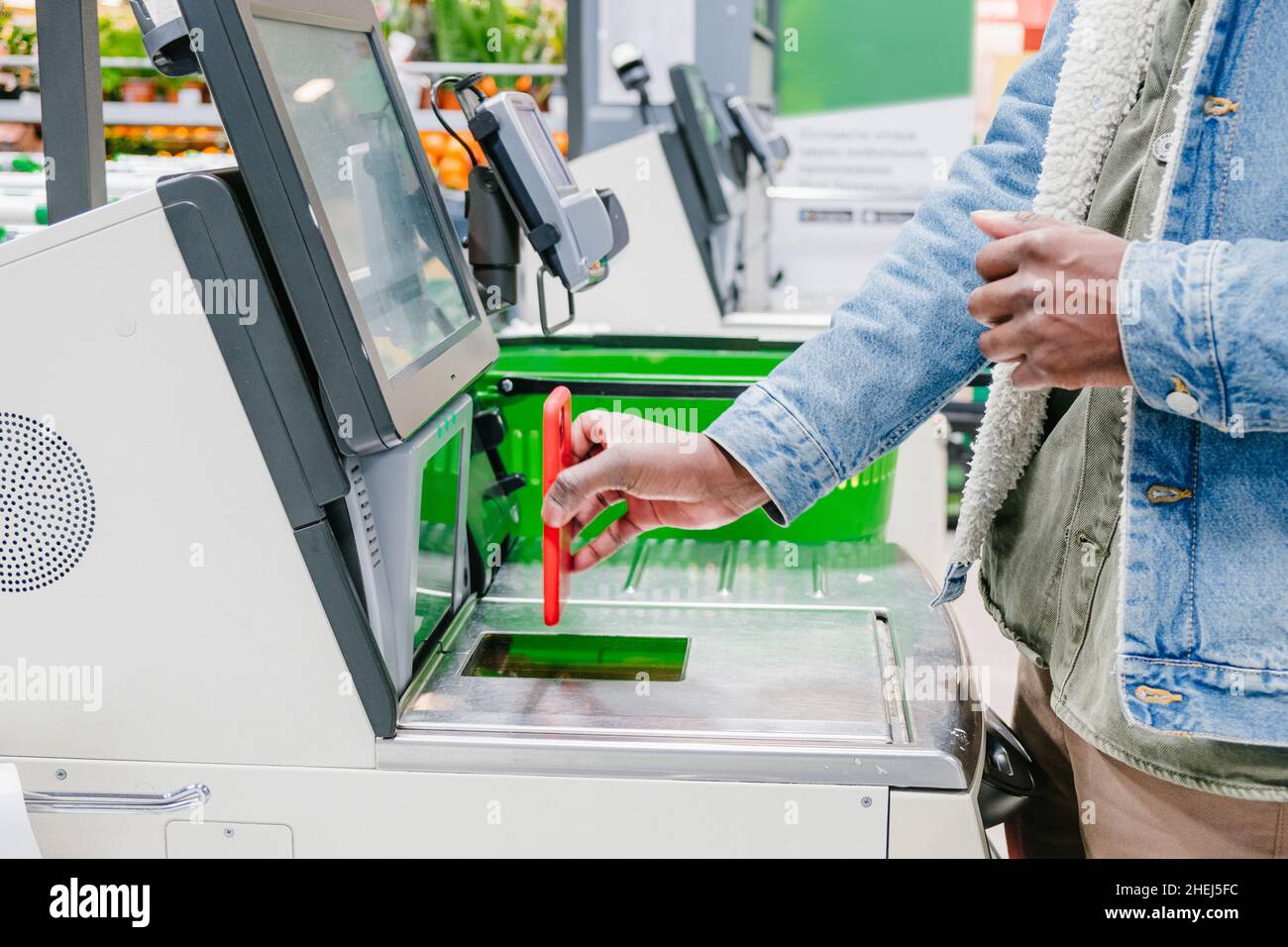 Homme noir dans une veste en denim chaud utilise un smartphone pour payer l'achat au point de caisse libre-service dans un supermarché à proximité Banque D'Images