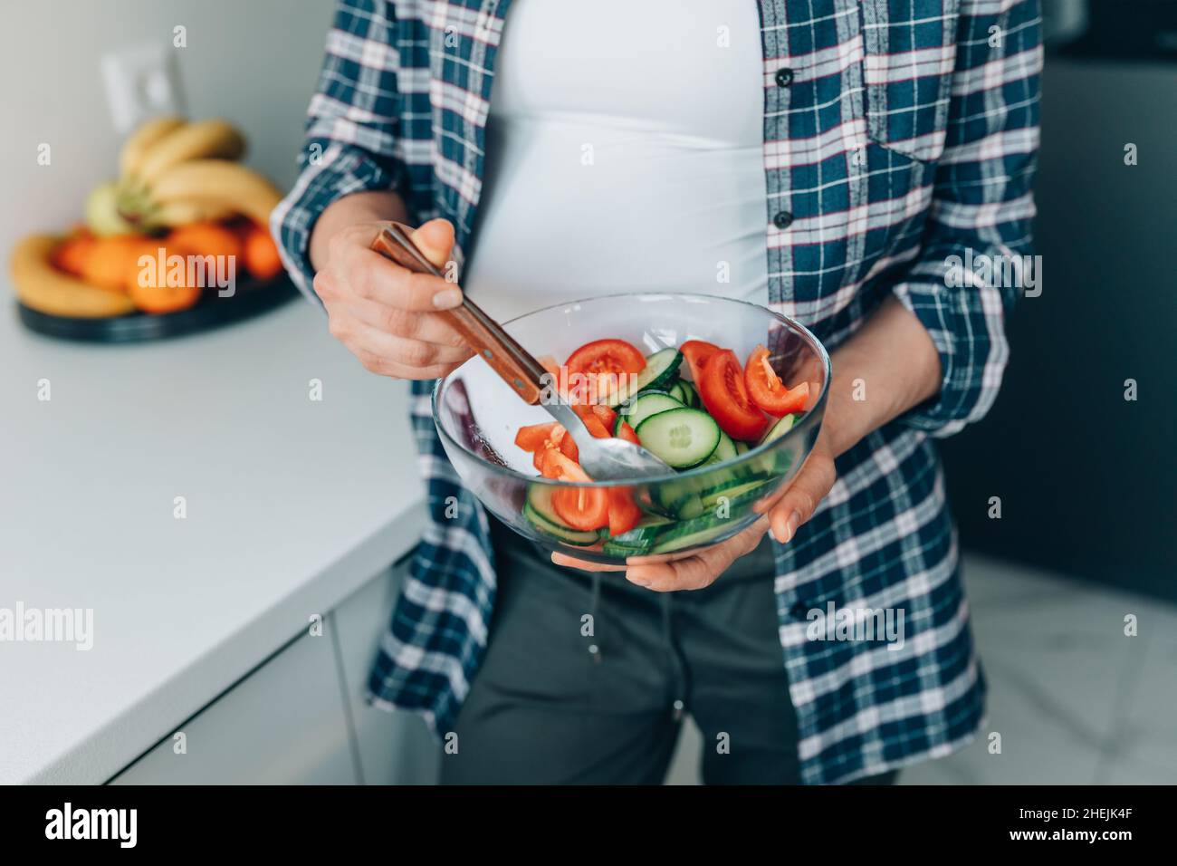 femme enceinte en haut blanc et chemise à carreaux mélanger la salade avec les concombres et les tomates dans le bol de la cuisine. Banque D'Images