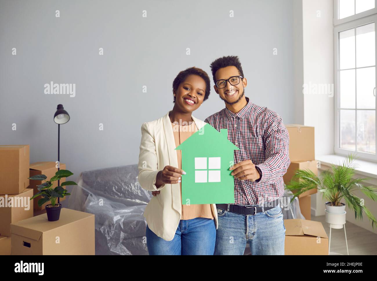 Joyeux jeune couple noir debout dans leur nouvelle maison, tenant la maison de papier et souriant Banque D'Images