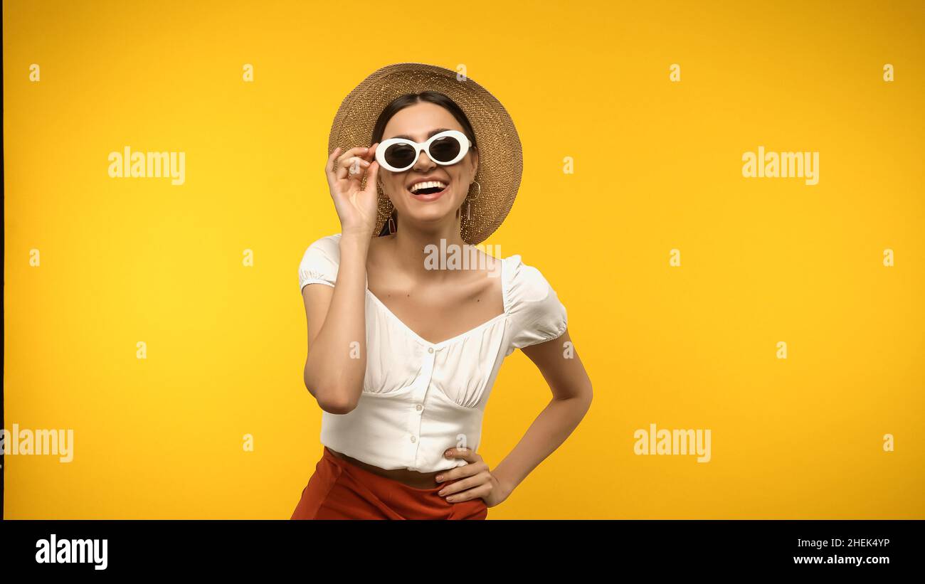 Femme gaie tenant des lunettes de soleil et main sur la hanche isolée sur jaune Banque D'Images