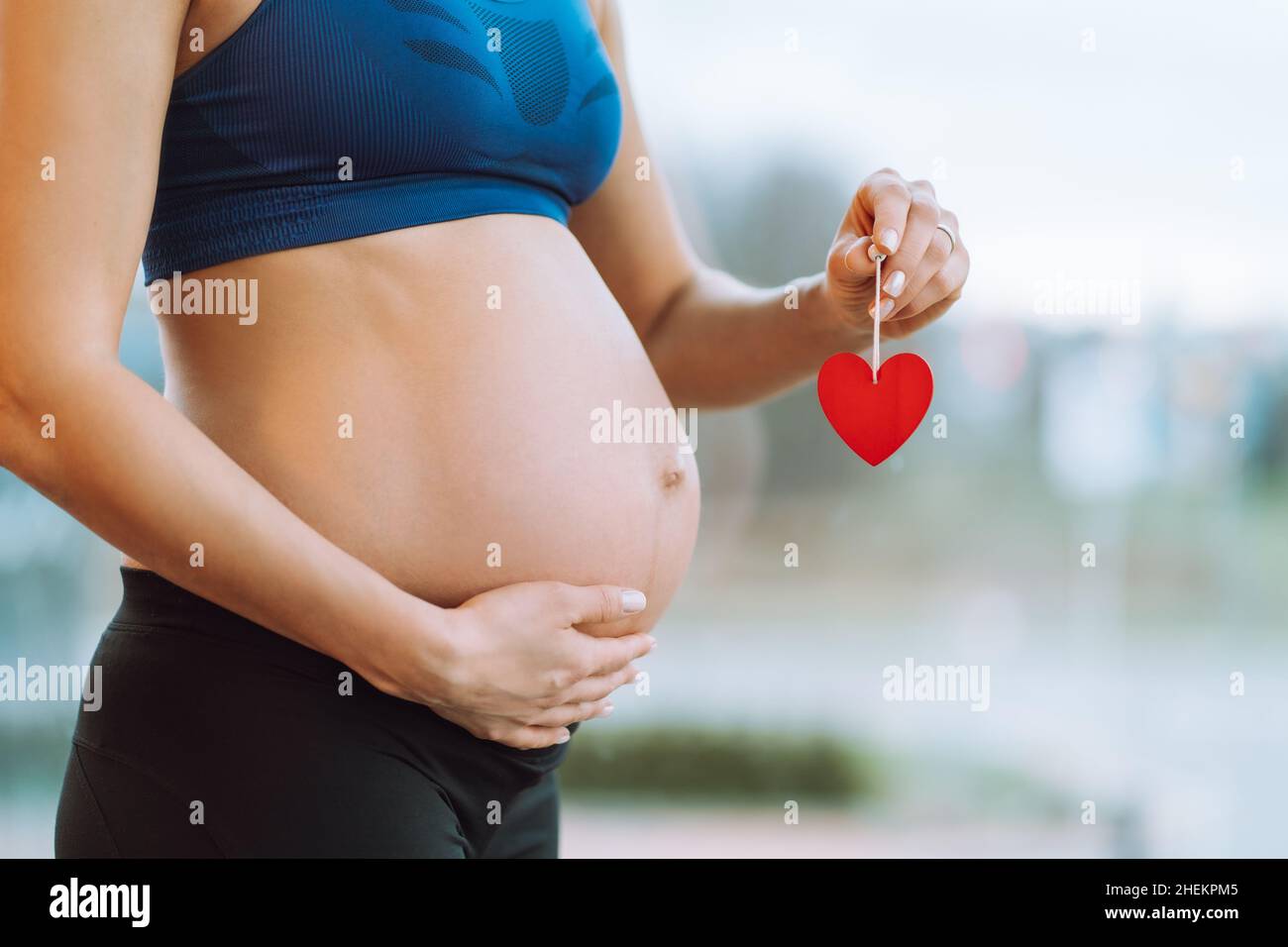 Jeune femme enceinte en vêtements de sport tenant le coeur rouge accroché à la main sur le bâton et caressant le ventre. Banque D'Images