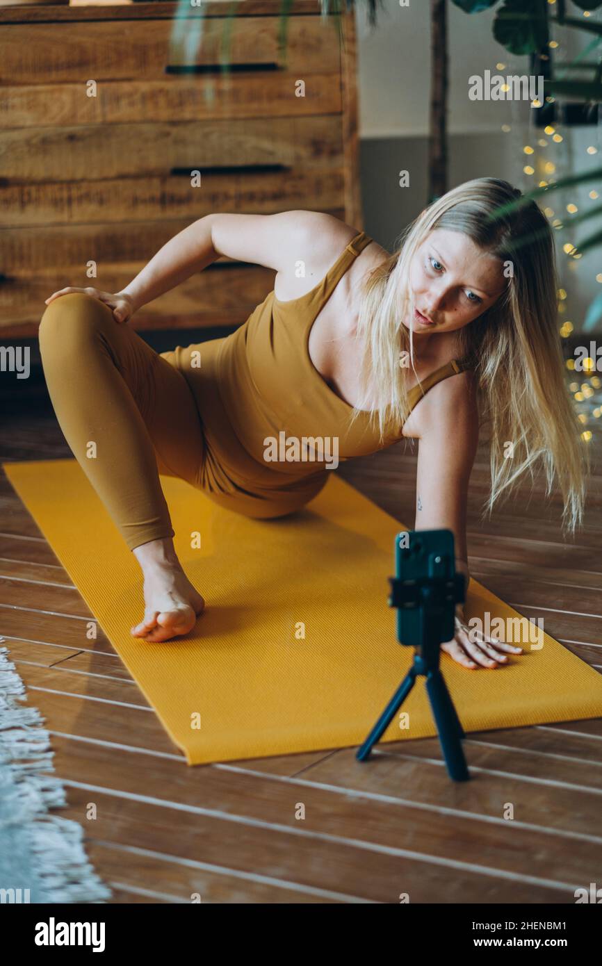 Femme avec de longs cheveux blond exercices sur le tapis avec grand sourire après la leçon en ligne par téléphone sur trépied debout sur le sol de parquet contre l'armoire Banque D'Images