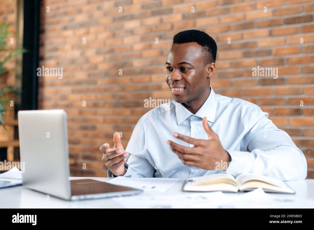 Afro-américain positif homme d'affaires réussi en chemise, pdg société, gestionnaire, courtier, utilise ordinateur portable pour la communication à distance avec le client, assis dans un bureau moderne, gestant avec ses mains, souriant Banque D'Images