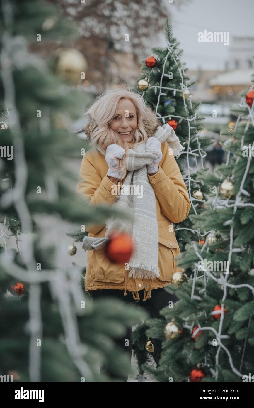 Jolie femme âgée aux cheveux blonds sur la place parmi les arbres de Noël. Banque D'Images