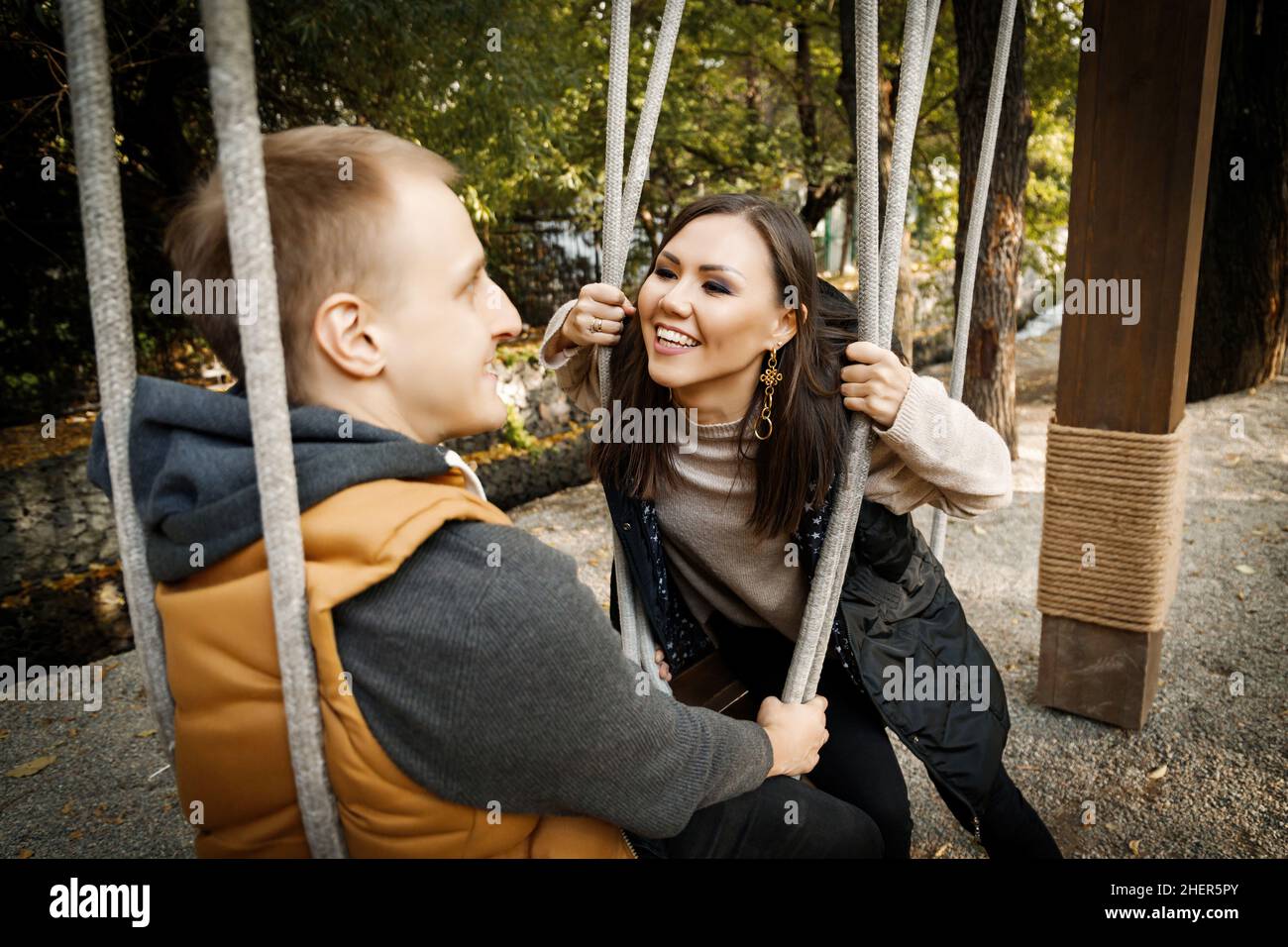Joyeux jeune couple de course mixte regardant l'un l'autre tout en s'asseyant sur une balançoire à l'extérieur. Banque D'Images