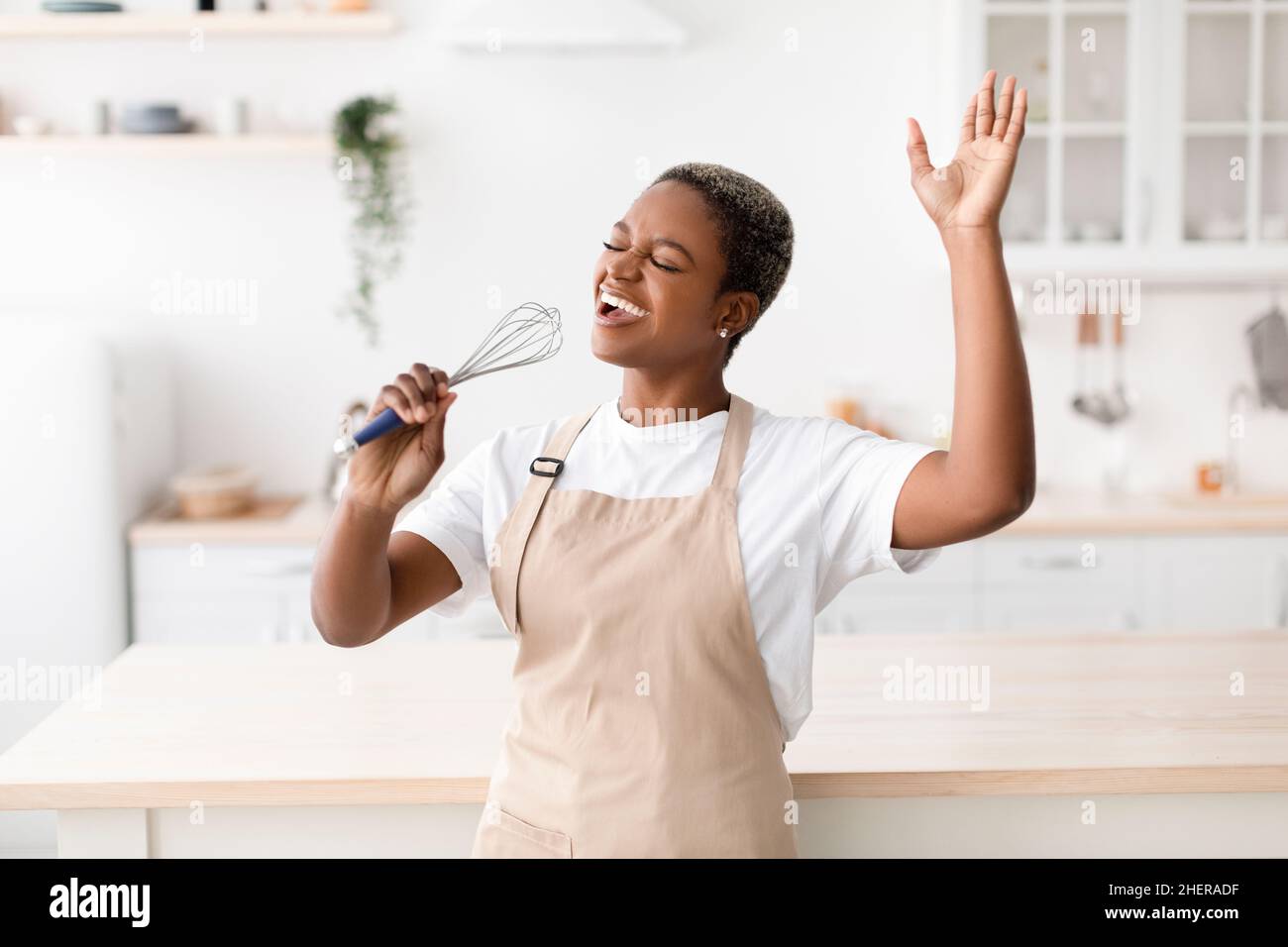 Une jeune femme afro-américaine, inspirée et heureuse, prépare de la nourriture, chante au microphone imaginaire Banque D'Images Une jeune femme afro-américaine, inspirée et heureuse, prépare de la nourriture, chante au microphone imaginaire Banque D'Images