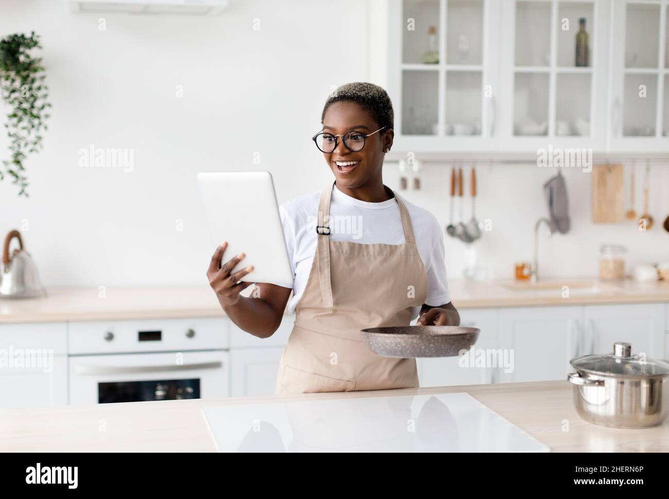 Surprise jeune africaine américaine heureuse femme en tablier et lunettes regarde la tablette et prépare le dîner Banque D'Images