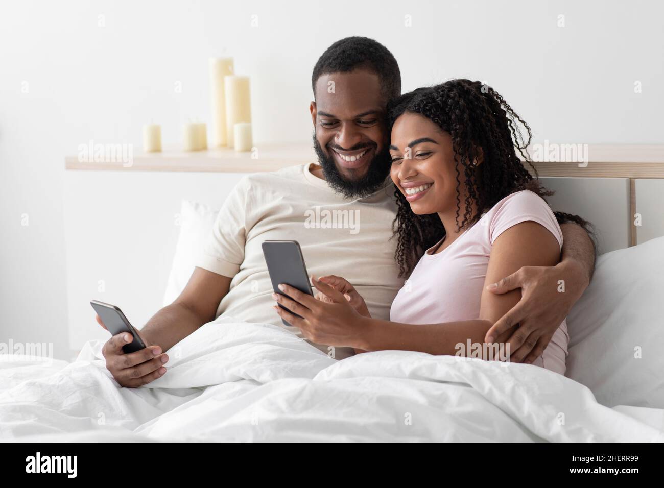Souriant, un jeune afro-américain câlins la dame et la dactylographie, le surf, le chat sur les smartphones sur un lit confortable Banque D'Images