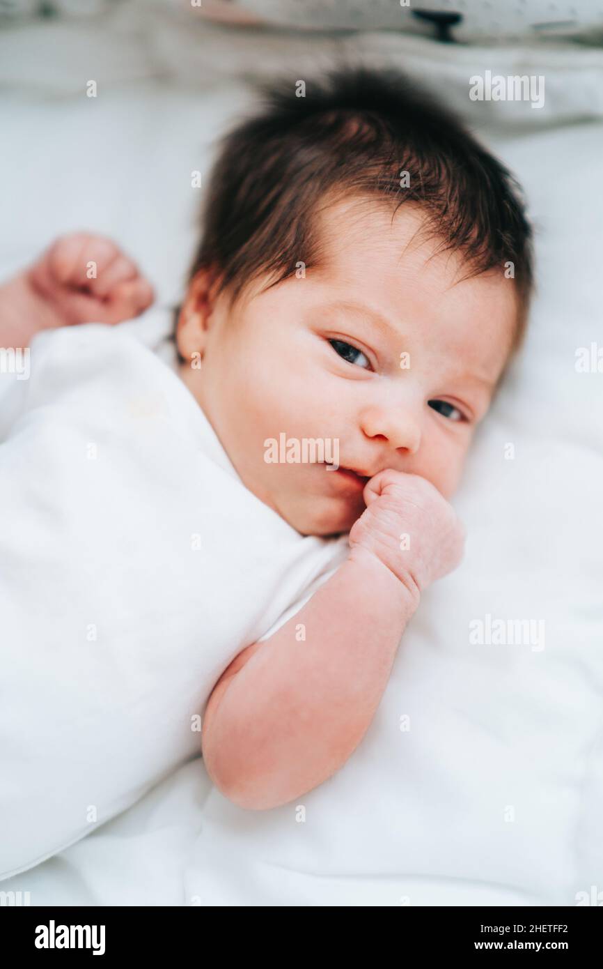 Mignon petit bébé nouveau-né avec de courts cheveux foncés dans le body blanc regarder endormi à la caméra clenching poings. Banque D'Images