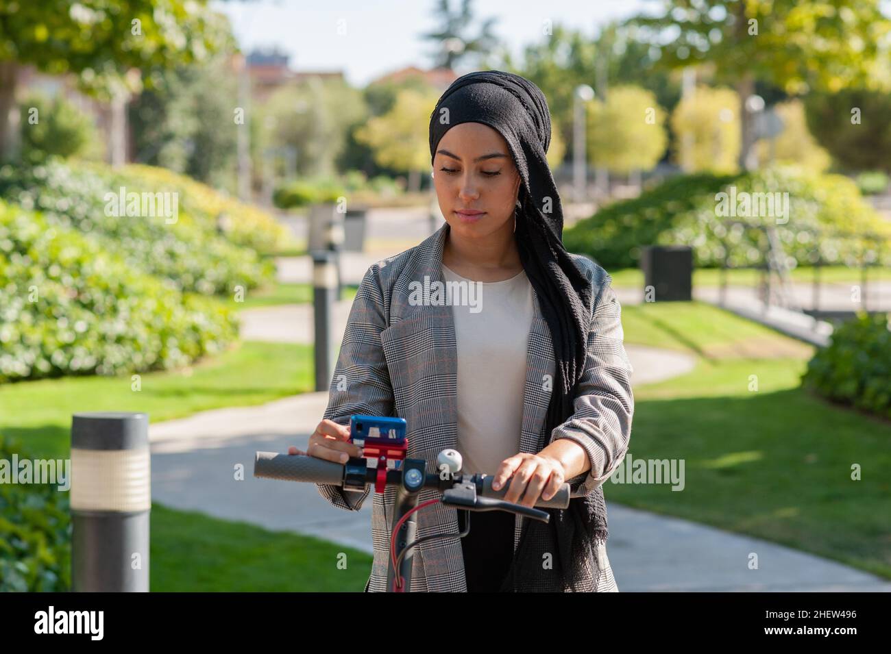 Vue de face d'une femme musulmane adulte utilisant son téléphone sur un scooter électrique dans le parc en une journée ensoleillée. Banque D'Images