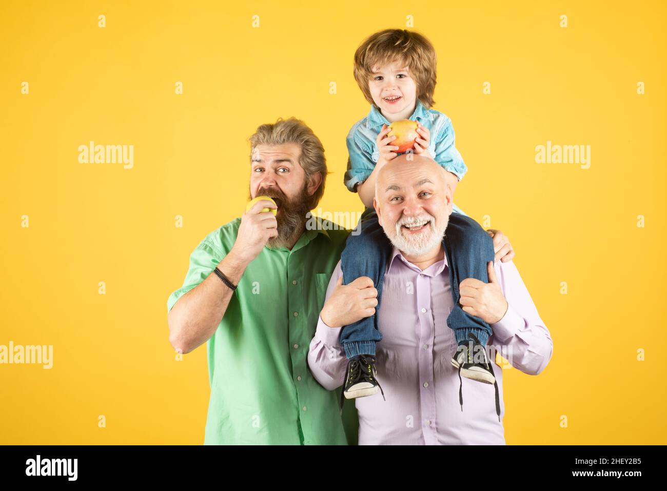Grand-père père et grand fils embrassant et mangeant de la pomme.Hommes de différents âges, isolés sur le jaune. Banque D'Images
