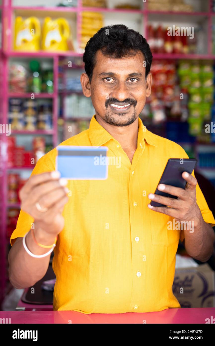 Kirana ou marchand d'épicerie montrant la carte de crédit à l'appareil photo après sur le téléphone mobile - concept d'accepter le paiement numérique dans les affaires de détail, cashless Banque D'Images