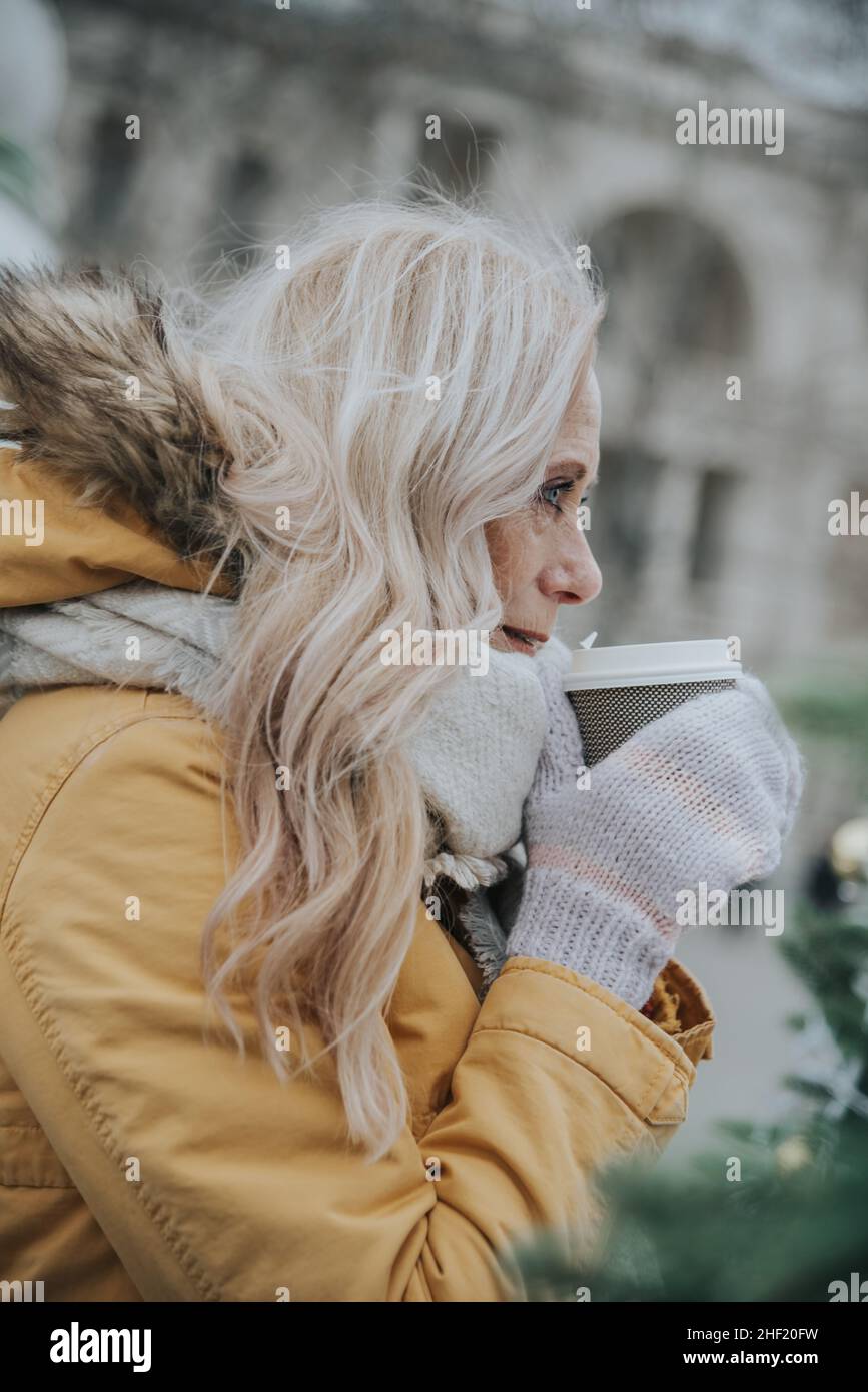 Une jolie femme âgée aux cheveux blonds boit du café sur la place parmi les arbres de Noël. Banque D'Images