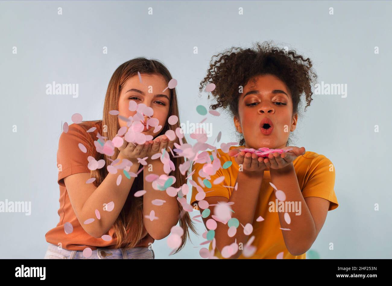 Deux joyeuses amies multiraciales qui soufflent des confettis à l'appareil photo et s'amusent tout en se tenant isolée sur fond gris, frien positif femelle Banque D'Images