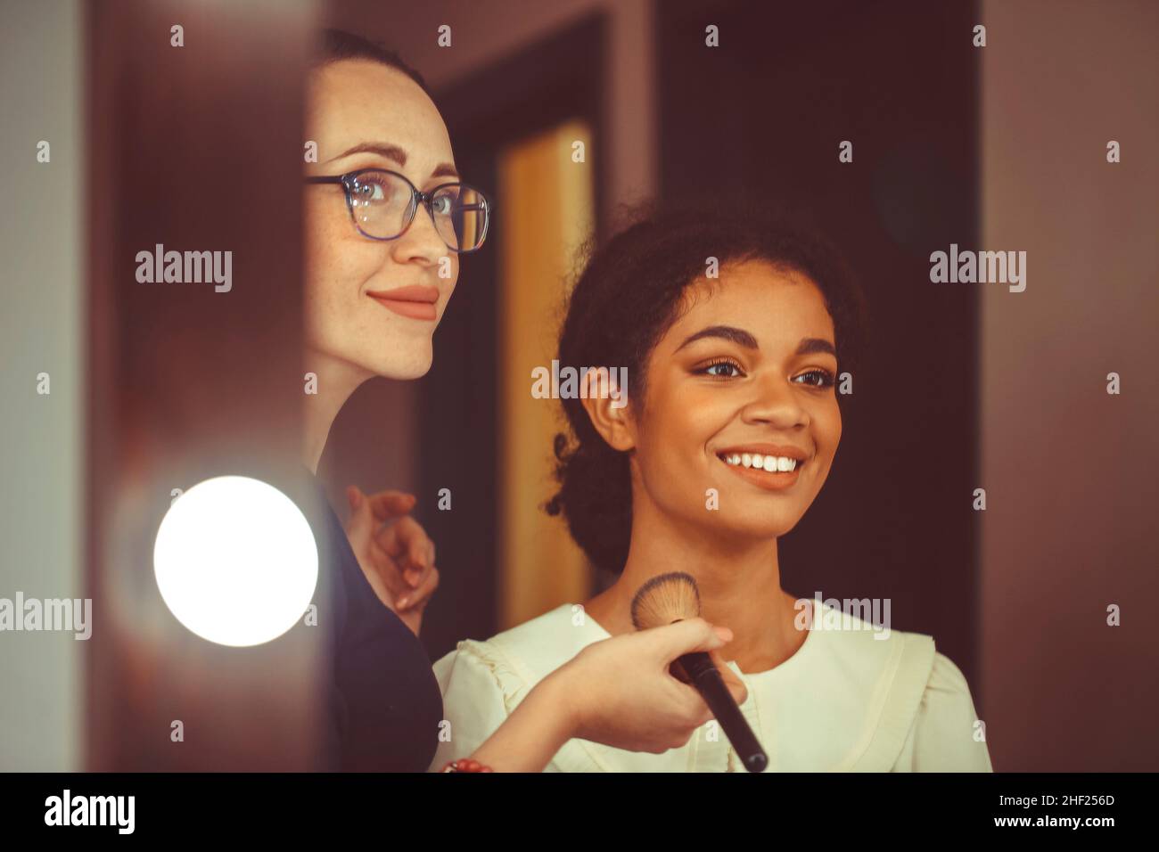 Jeune heureuse afro-américaine attrayante femme rendant visite à un maquilleur dans un salon de beauté, visagiste profesoional tenant la brosse appliquant le maquillage sur le visage de Banque D'Images