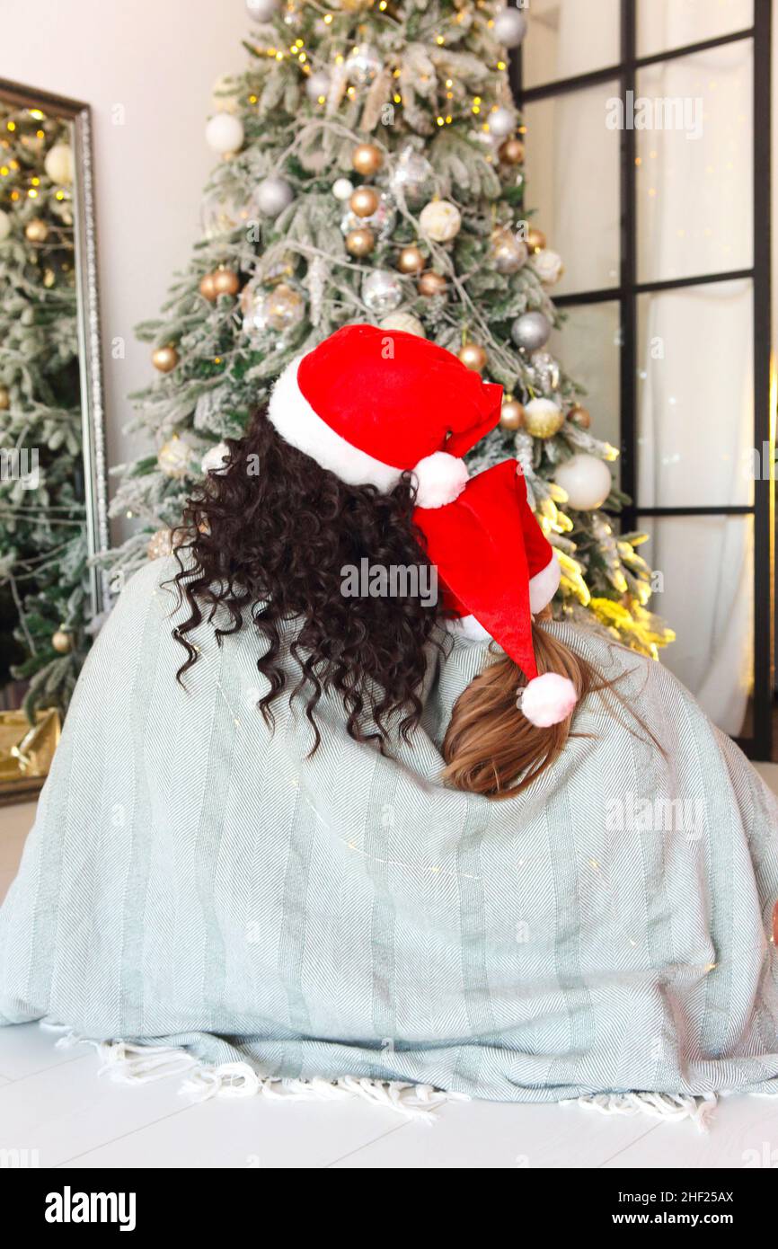 Noël en famille.Vue arrière de la mère et de l'enfant portant des chapeaux de père noël assis sous une couverture tricotée sur le sol et embrassant, en regardant l'arbre de Noël décoré. Banque D'Images