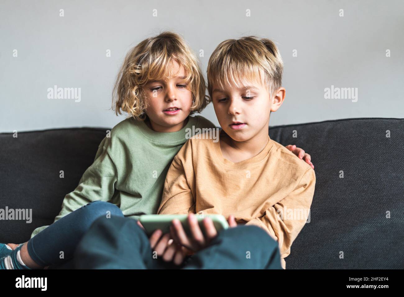 Adorable petite sœur et frère aux cheveux blonds dans des vêtements décontractés qui se câblent tout en étant assis sur un canapé et en regardant une vidéo intéressante sur un téléphone portable Banque D'Images