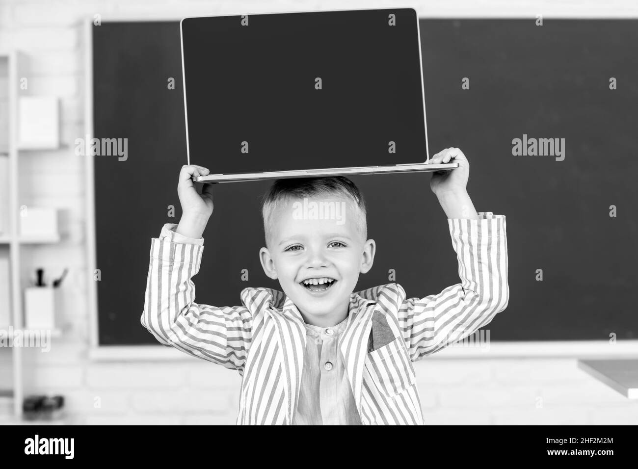 Petit garçon étudiant tenant un ordinateur portable à la tête en classe Banque D'Images