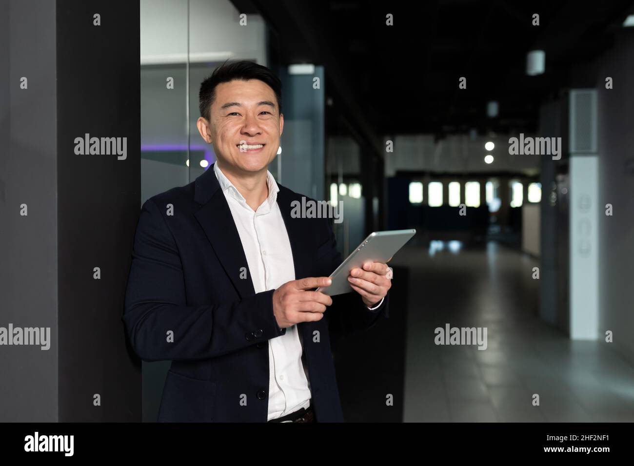 Un homme d'affaires asiatique prospère, le patron, dans un costume d'affaires se réjouit et sourit utilise une tablette, travaille dans un bureau moderne Banque D'Images